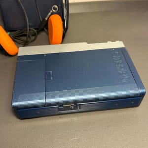 May include: A blue and silver portable cassette player with the word STEREO embossed on the front. The device features a side-mounted volume dial and is accompanied by a pair of black headphones with bright orange foam ear pads and a cord.