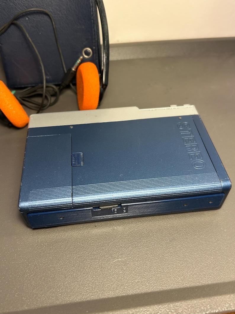 May include: A blue and silver portable cassette player with the word STEREO embossed on the front. The device features a side-mounted volume dial and is accompanied by a pair of black headphones with bright orange foam ear pads and a cord.