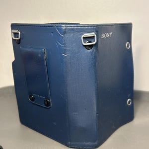 May include: A dark blue Sony camera case made of a textured synthetic material. The case features a vertical belt clip on the back, two silver-toned metal loops for a shoulder strap, and two small circular metal grommets on the right side panel.