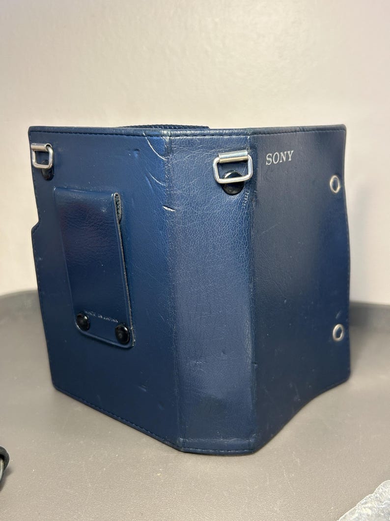 May include: A dark blue Sony camera case made of a textured synthetic material. The case features a vertical belt clip on the back, two silver-toned metal loops for a shoulder strap, and two small circular metal grommets on the right side panel.