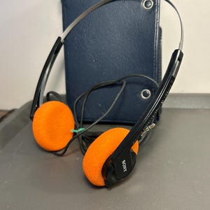 May include: A pair of black Sony headphones with bright orange foam ear pads and a thin metal headband. The headphones feature a black cord and are positioned in front of a dark blue, rectangular Sony branded case with two metal snap closures.