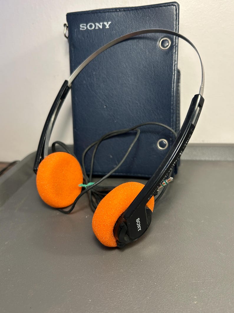 May include: A pair of black Sony headphones with bright orange foam ear pads and a thin metal headband. The headphones feature a black cord and are positioned in front of a dark blue, rectangular Sony branded case with two metal snap closures.