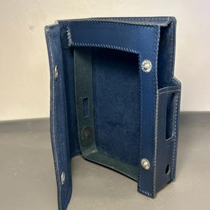 May include: A dark blue leather protective case with a hinged flap, featuring visible stitching along the edges, silver-toned metal snap fasteners, and precise cutouts on the side for device access, designed to hold a rectangular electronic device.