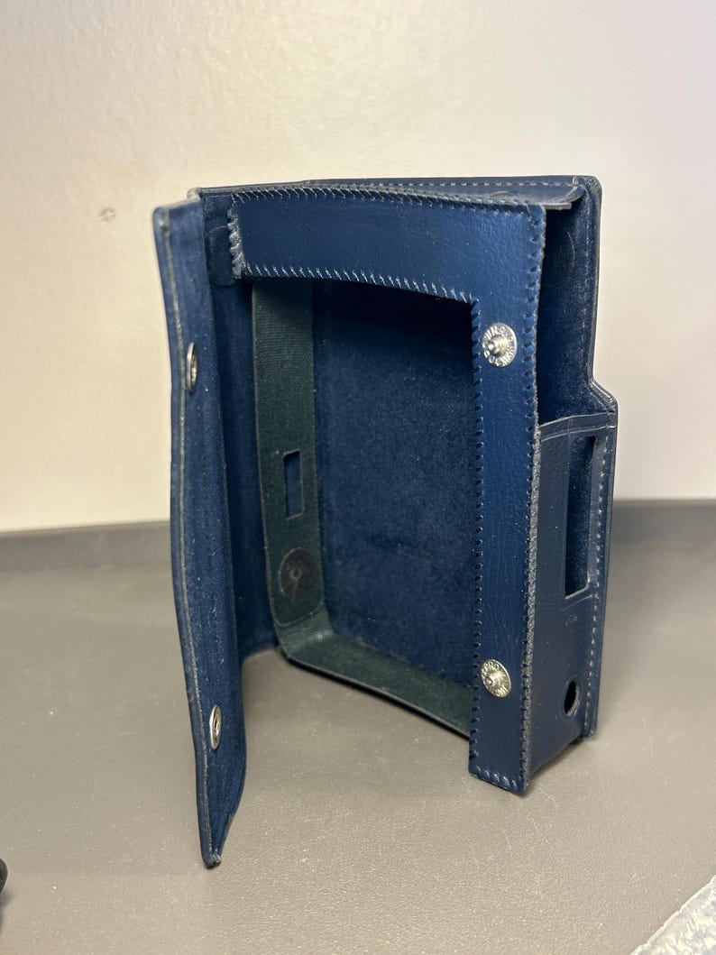 May include: A dark blue leather protective case with a hinged flap, featuring visible stitching along the edges, silver-toned metal snap fasteners, and precise cutouts on the side for device access, designed to hold a rectangular electronic device.