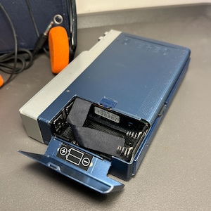 May include: A blue and silver portable cassette player with an open battery compartment. The interior shows a black ribbon and a white label with the text SERIAL NO. 108287. The battery door features a diagram indicating positive and negative polarity.