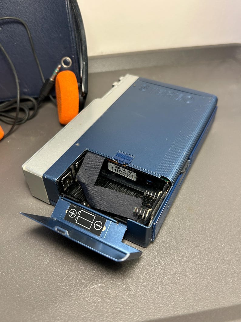 May include: A blue and silver portable cassette player with an open battery compartment. The interior shows a black ribbon and a white label with the text SERIAL NO. 108287. The battery door features a diagram indicating positive and negative polarity.