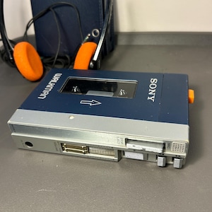 May include: A blue and silver Sony Walkman cassette player with a clear window, featuring volume sliders, playback control buttons, and a microphone port. The device is accompanied by a pair of black headphones with bright orange foam ear pads.