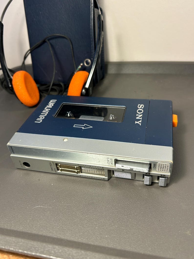 May include: A blue and silver Sony Walkman cassette player with a clear window, featuring volume sliders, playback control buttons, and a microphone port. The device is accompanied by a pair of black headphones with bright orange foam ear pads.