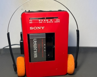 May include: A red Sony Walkman FM/AM radio cassette player with orange ear pads. The device has a silver headband and black earphone components. The Walkman has the text "WALKMAN FM/AM" and "SONY" printed on the front.