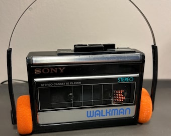 May include: A black Sony Walkman stereo cassette player with a silver front panel and the text "STEREO CASSETTE PLAYER" and "WALKMAN" printed on it. The device is attached to a thin metal headband with bright orange foam ear pads on each side.
