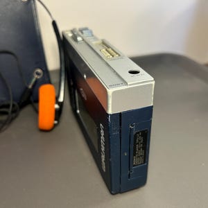 May include: A dark blue Sony Walkman TPS-L2 cassette player with a silver top panel featuring volume controls and a DC input. The device includes a black battery compartment cover and is accompanied by a pair of headphones with orange foam pads.