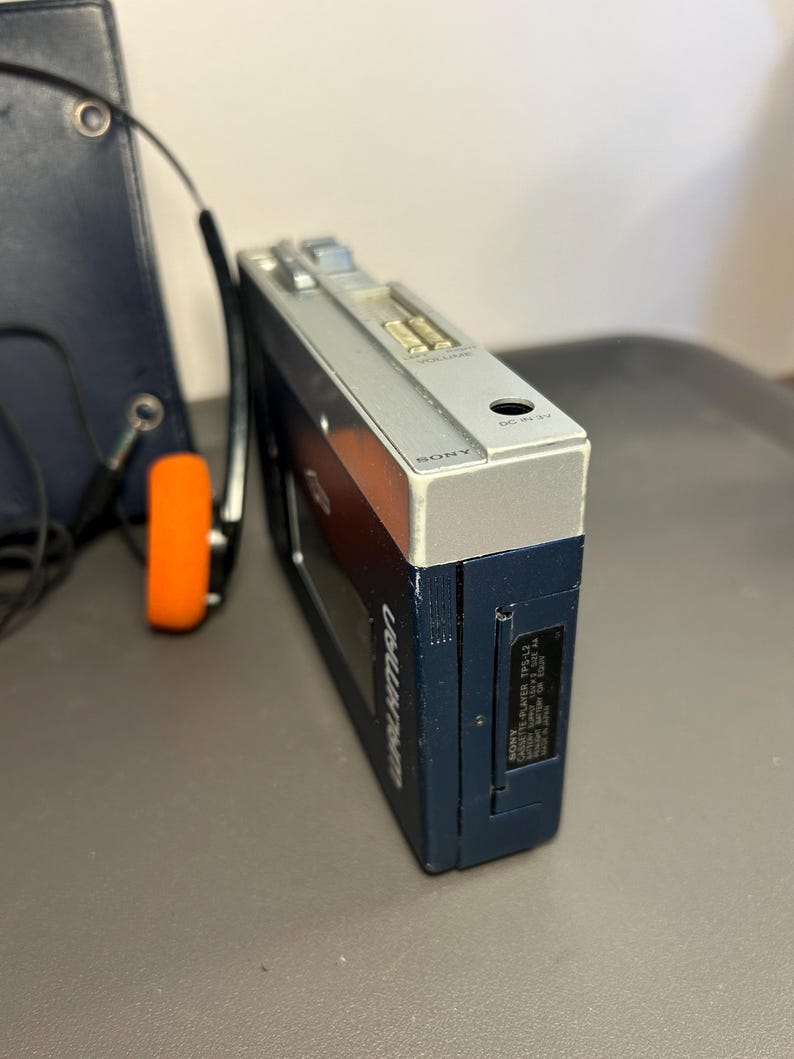 May include: A dark blue Sony Walkman TPS-L2 cassette player with a silver top panel featuring volume controls and a DC input. The device includes a black battery compartment cover and is accompanied by a pair of headphones with orange foam pads.