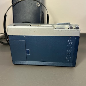 May include: A blue and silver portable cassette player with the word STEREO embossed on the side. The device features top-mounted control buttons and a headphone jack. A pair of black headphones and a dark blue Sony branded carrying case are visible.