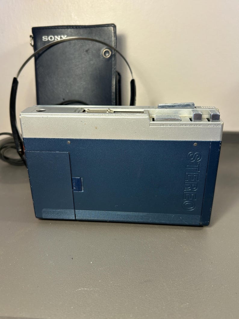 May include: A blue and silver portable cassette player with the word STEREO embossed on the side. The device features top-mounted control buttons and a headphone jack. A pair of black headphones and a dark blue Sony branded carrying case are visible.