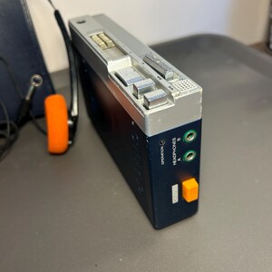 May include: A portable cassette player with a dark blue body and a silver top panel. It features two green headphone jacks labeled A and B, an orange button, and silver control switches. A pair of black headphones with orange foam ear pads is attached.