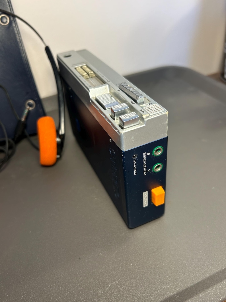 May include: A portable cassette player with a dark blue body and a silver top panel. It features two green headphone jacks labeled A and B, an orange button, and silver control switches. A pair of black headphones with orange foam ear pads is attached.