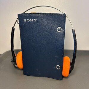 May include: A dark blue Sony portable cassette player encased in a leather-like cover with a silver metal snap closure. The device features a thin metal headband arching over the top, connected to black earphone arms with bright orange foam pads.