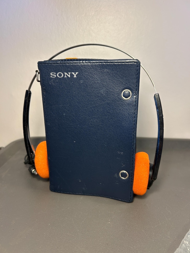 May include: A dark blue Sony portable cassette player encased in a leather-like cover with a silver metal snap closure. The device features a thin metal headband arching over the top, connected to black earphone arms with bright orange foam pads.