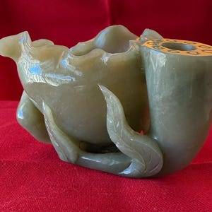 May include: A green jade carving of a lotus flower with a hollow center. The flower is carved with intricate details and has a long stem.