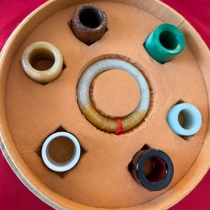 Könnte beinhalten: Eine runde Box mit beigefarbenem Innenraum enthält eine Sammlung von Jade-Schmuck. Das Set umfasst ein Jade-Armreif und mehrere Jade-Ringe in verschiedenen Farben, darunter Weiß, Grün und Schwarz. Die Box befindet sich vor einem roten Hintergrund.
