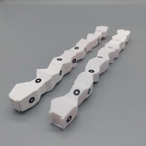 Puede incluir: Dos juguetes blancos segmentados con forma de serpiente con marcas circulares negras. Los juguetes tienen un diseño geométrico y están colocados en diagonal sobre un fondo gris. Los juguetes parecen ser de plástico.