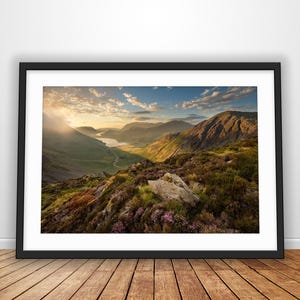 May include: A framed landscape photograph featuring a mountain valley at sunset with a lake in the distance, purple heather in the foreground, and a bright sky with scattered clouds, all presented in a black frame with a white mat border.