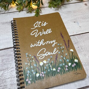 May include: Spiral-bound notebook with a brown cover. The cover features the white text "It is Well with my Soul" above a painted floral design with blue, white, and pink flowers. The artist's signature "Tiah" is in the bottom right corner.