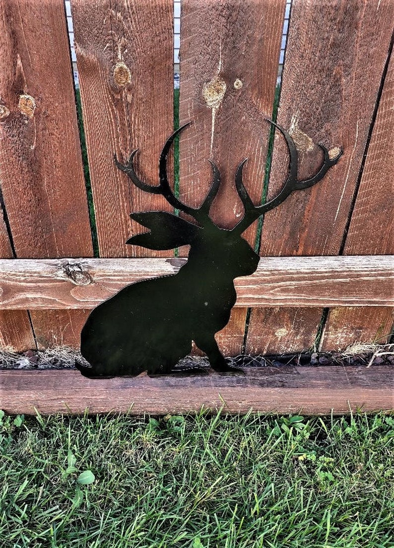 May include: Black metal silhouette of a rabbit with large antlers, sitting on a wooden fence post in front of a wooden fence. The rabbit is facing to the right.