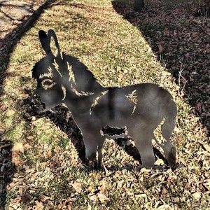 May include: A black metal silhouette of a donkey standing on a bed of brown leaves. The donkey is facing the left side of the image.