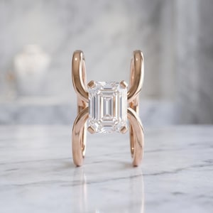May include: A rose gold ring featuring a central emerald-cut clear gemstone held by four prongs. The band consists of two curved, parallel metal segments that arch upwards and downwards from the centre stone, creating an open, symmetrical design.