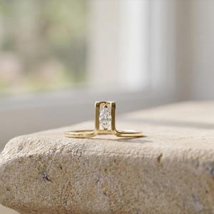 Peut inclure: Une bague en or au design unique, ornée de deux diamants taille marquise disposés verticalement dans un cadre rectangulaire. La monture de la bague s'étend de chaque côté, créant un look moderne et élégant. La bague est présentée sur une surface en pierre claire.