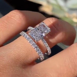 Könnte beinhalten: Ein silberner Verlobungsring mit einem großen, rechteckigen, klaren Diamanten. Der Ring hat eine Band aus kleineren Diamanten.