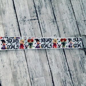 Puede incluir: Cordón blanco con un patrón repetido de brujas de dibujos animados y la frase "SQUAD GOALS" en negro. Tiene un cierre y un llavero de metal plateado.