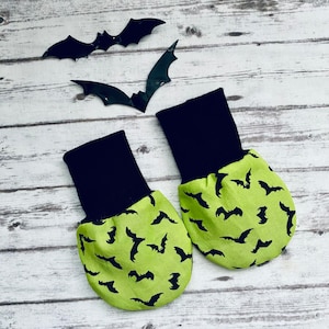 Może przedstawiać: Para bucików dla niemowląt z motywem Halloween. Buciki mają czarny mankiet i limonkową stopę z powtarzającym się czarnym wzorem nietoperzy. Trzy czarne dekoracje nietoperzy umieszczono nad bucikami na białej drewnianej powierzchni.