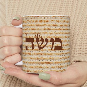May include: White ceramic mug featuring a matzah pattern and the Hebrew word "Moshe" in brown. The mug is held by a person wearing a beige sweater. The design is a nod to Jewish traditions.