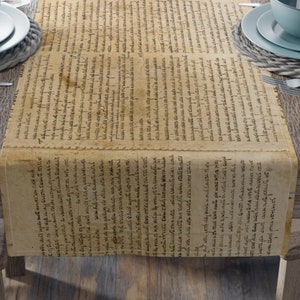 May include: A brown table runner with a vintage, handwritten text pattern. The runner is set on a wooden table with two place settings on each side. Each place setting includes a blue bowl, a white napkin, a wine glass, and silverware.