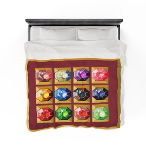 K&ouml;nnte beinhalten: Eine Bettdecke mit einem sich wiederholenden Muster aus bunten Edelsteinen. Jeder Edelstein ist in einem goldenen Quadrat auf einem weinroten Hintergrund gesetzt. Die Bettdecke liegt auf einem Bett mit wei&szlig;en Laken und Kissen.