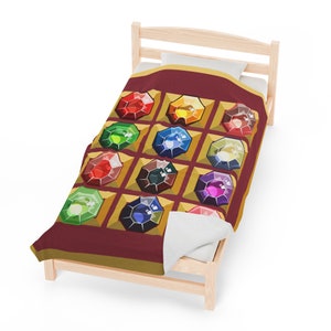 K&ouml;nnte beinhalten: Eine weinrote Decke mit einem sich wiederholenden Muster aus bunten, juwelenartigen Formen in goldenen Rahmen. Die Decke wird auf einem kleinen, hellen Holzbetttisch mit einem wei&szlig;en Kissen pr&auml;sentiert.