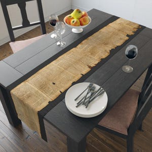 May include: A dark wood dining table set for a meal. A long, parchment-style table runner with text runs down the center. A white bowl of apples sits at the top. Wine glasses, plates, and silverware complete the setting.