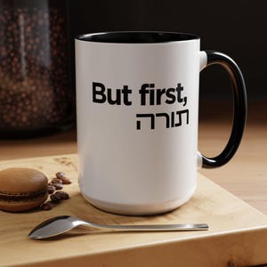 May include: White ceramic mug with a black interior and handle. The mug features the text "But first, תורה" in black. A spoon, coffee beans, and a macaron are on a wooden surface.