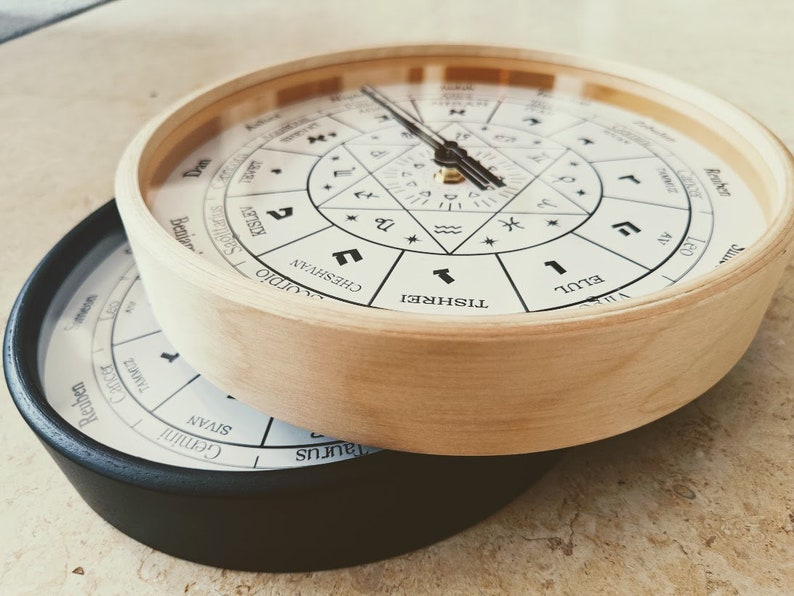 May include: Two round wooden clocks with black trim. The top clock has a white face with Hebrew text and symbols. The bottom clock has a black face with Hebrew text and symbols.