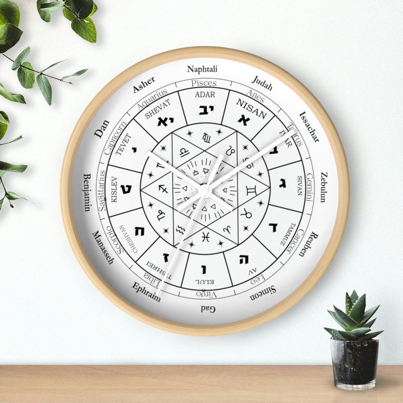 May include: A white circular clock with a wooden frame. The clock face features a black and white design with Hebrew text and astrological symbols. The clock hands are white.