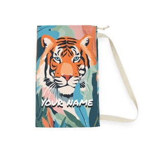 Puede incluir: Una bolsa con cordón con una ilustración de tigre y el texto "YOUR NAME". El tigre tiene rayas naranjas y negras y ojos azules, sobre un fondo de follaje colorido. La bolsa tiene una correa color crema.