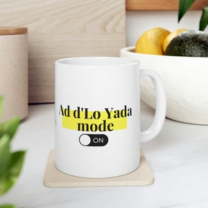 Puede incluir: Taza de cerámica blanca con el texto "Ad d'Lo Yada mode ON" en letras negras sobre un rectángulo amarillo. La taza está sobre un posavasos beige. La taza tiene un asa blanca.