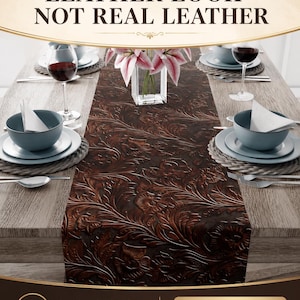 May include: A table setting with a brown leather-look polyester fabric table runner. The runner features a floral embossed design. The table is set with blue bowls, white plates, wine glasses, and a vase of pink flowers. The text reads: LEATHER LOOK - NOT REAL LEATHER.