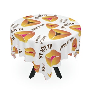 Puede incluir: Un mantel redondo blanco con un patrón repetido de pasteles de hamantaschen en tonos naranja, amarillo y rosa. Las palabras "Happy Purim" y texto hebreo también están impresas en la tela.