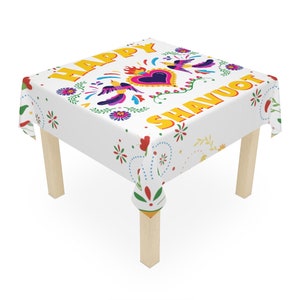 Puede incluir: Un mantel cuadrado blanco con un diseño floral y de pájaros colorido. Las palabras "HAPPY SHAVUOT" están impresas en amarillo. El mantel está sobre una pequeña mesa de madera.