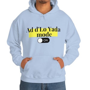 Puede incluir: Sudadera con capucha azul claro con el texto "Ad d'Lo Yada mode ON" en letras negras sobre un rectángulo amarillo. La sudadera tiene una capucha con cordón y un bolsillo delantero. El interruptor "ON" es blanco.