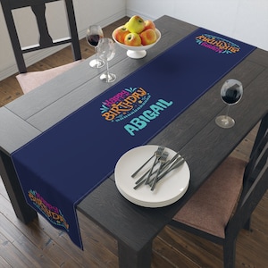 May include: A blue table runner with the text "Happy Birthday" in colorful letters and the name "Abigail" in white letters. The runner is on a wooden table with a plate of apples, two wine glasses, and a plate of silverware.