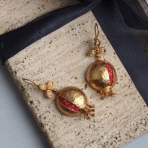 Puede incluir: Un par de pendientes de oro con forma de granadas con detalles de piedras preciosas rojas. Los pendientes se muestran sobre una superficie texturizada con una cinta negra.